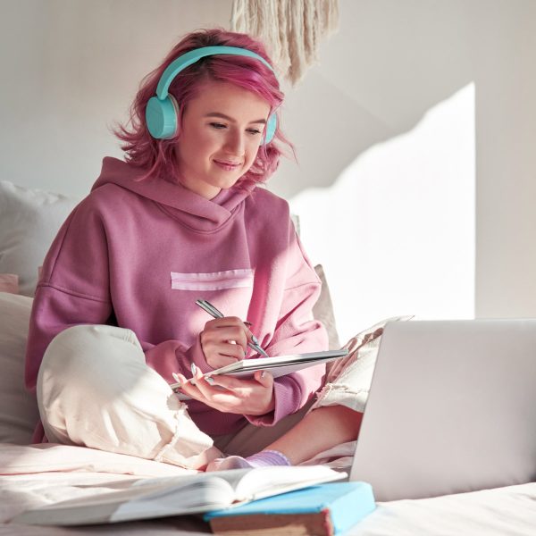 In einem hellen Raum sitzt eine junge Frau mit Kopfhörern im Schneidersitz vor einem aufgeklapptem Laptop und Büchern und macht sich Notizen.
